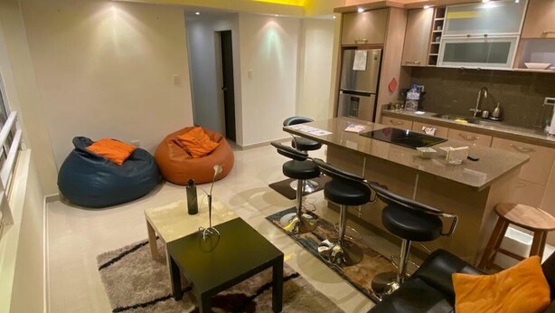 Una sala moderna con un sofá, un reposapiés y taburetes de bar. Hay una zona de cocina con un refrigerador, un microondas y un lavamanos.