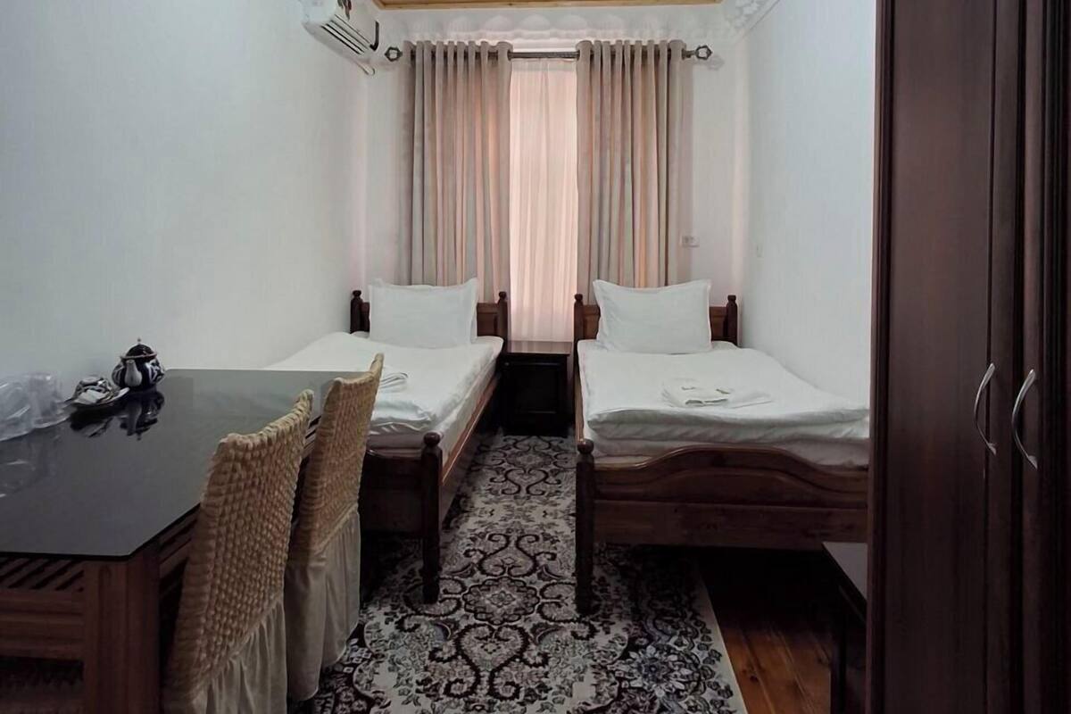 A room with two single beds, a wooden ceiling, a patterned rug, a table with chairs, and a wardrobe.