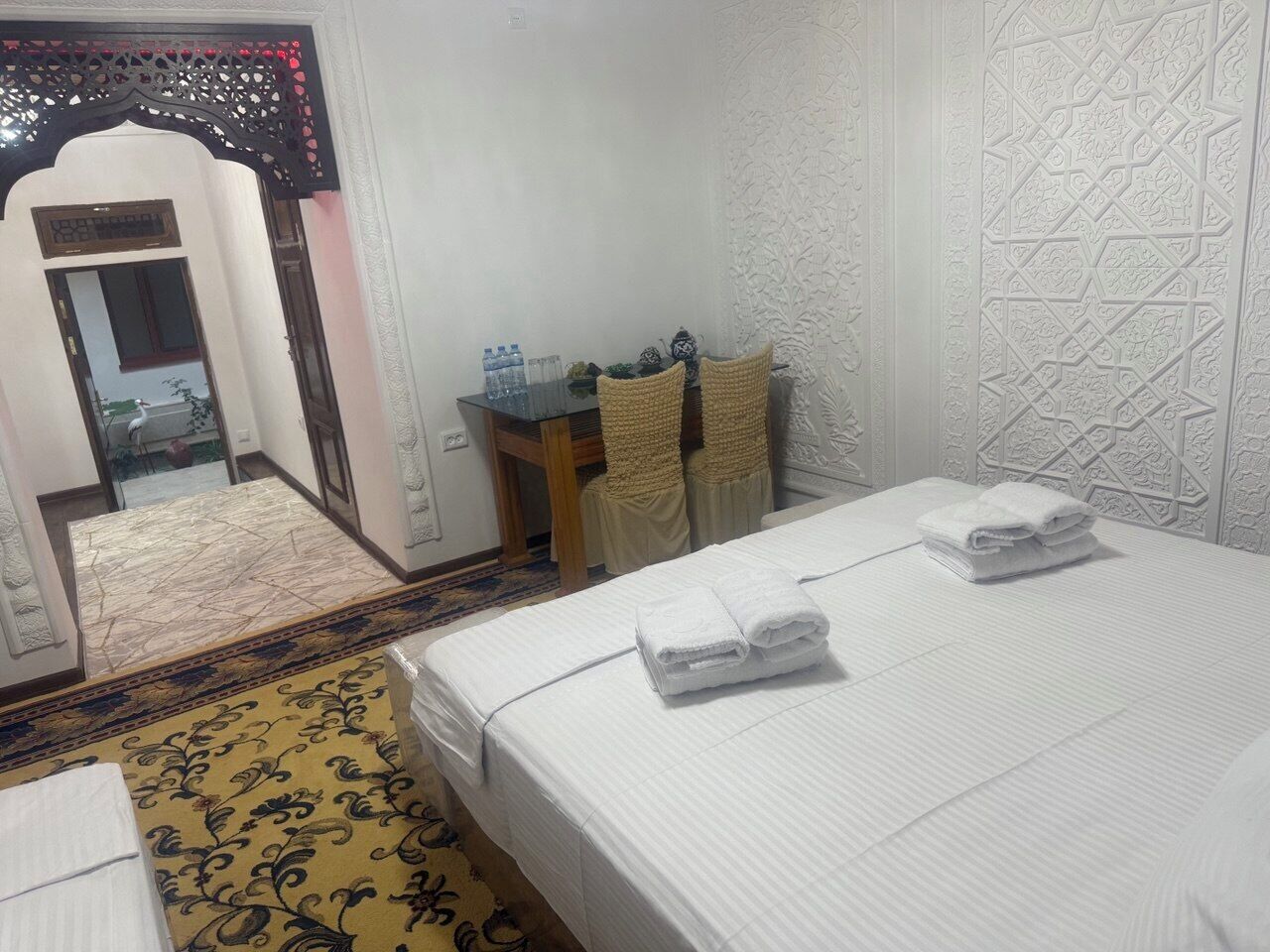 A hotel room with a bed, a small table with chairs, a decorative wall, and a patterned rug.