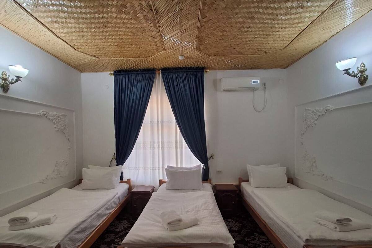 A room with two single beds, a large window with blue curtains, a woven ceiling, and a wall-mounted air conditioner.