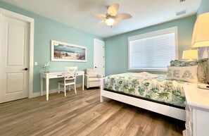 Ein Schlafzimmer mit Bett, Schreibtisch, Stuhl, Deckenventilator und einem Fenster mit Jalousien.