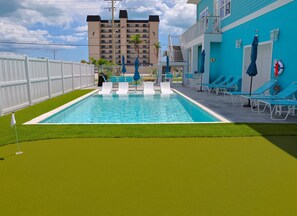 Ein Swimmingpool mit weißen Liegestühlen und Sonnenschirmen, umgeben von einem weißen Zaun und einem Gebäude mit blauer Fassade.