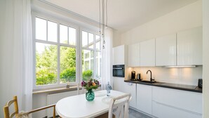Une cuisine moderne avec des meubles blancs, une table à manger avec des chaises, une fenêtre avec des rideaux et une vue sur la verdure à l’extérieur.