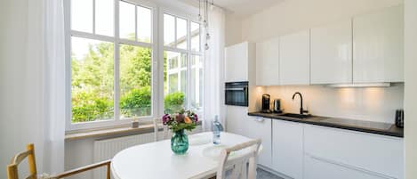 A modern kitchen with white cabinets, a dining table with chairs, a window with curtains, and a view of greenery outside.