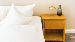 Un lit bien fait, avec du linge de lit blanc et des oreillers, une table de chevet en bois avec une lampe et un vase, et un sol recouvert d’une moquette.