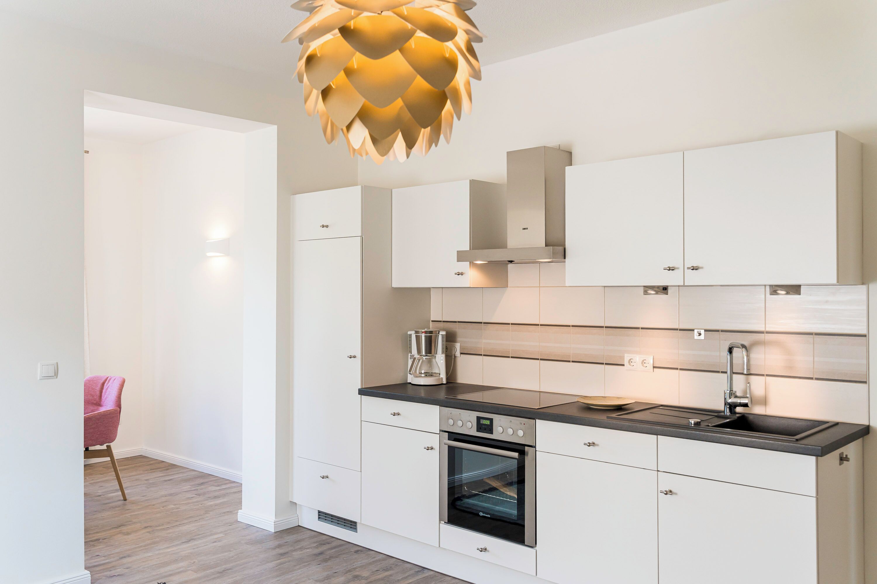 A modern kitchen with white cabinetry, a built-in oven, a stainless steel sink, and a unique pendant light.