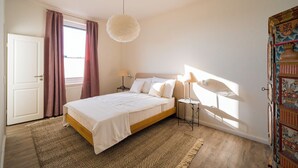 Un dormitorio con cama, lámparas de noche, una lámpara colgante, una ventana con cortinas y una puerta.