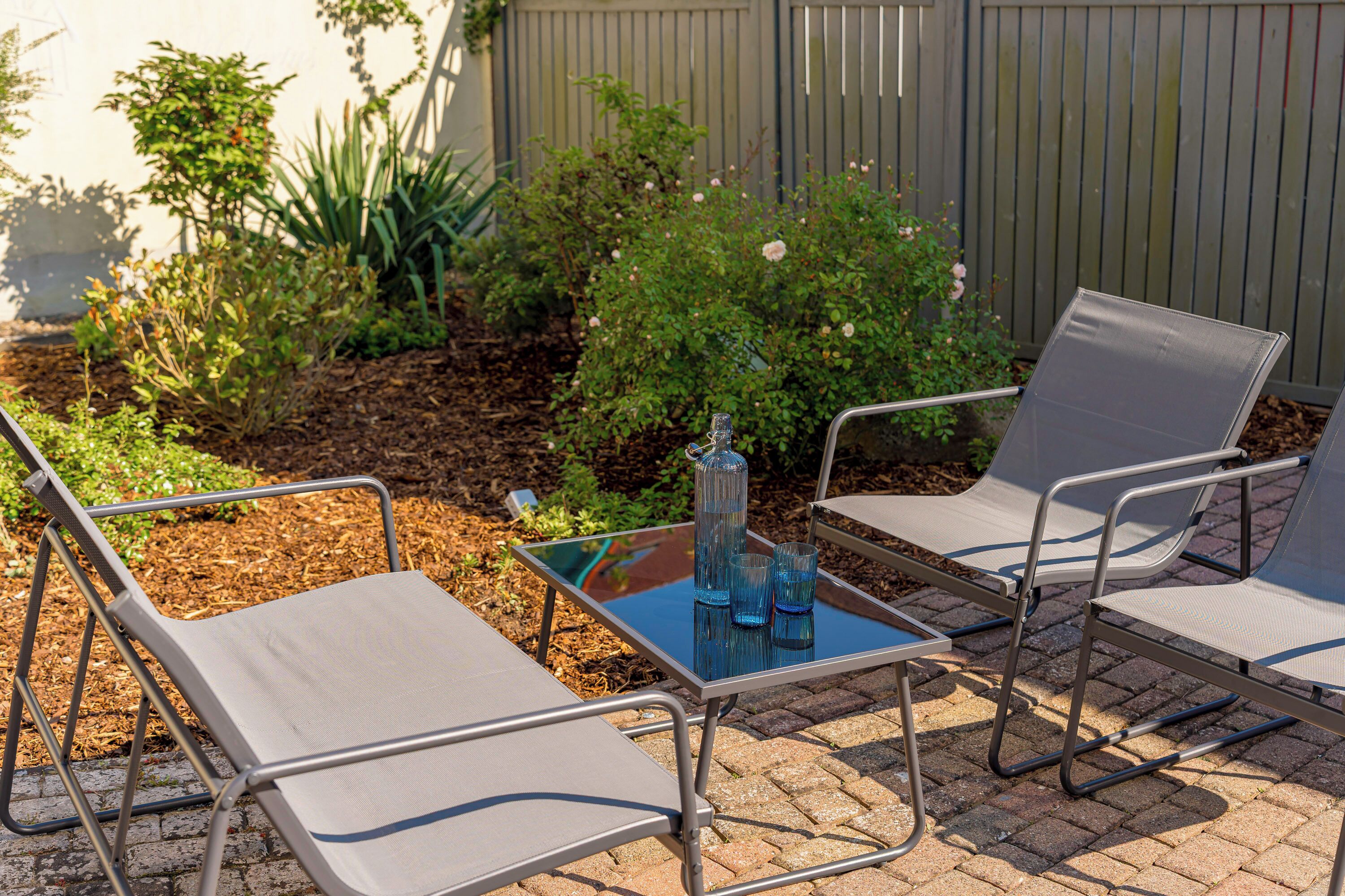 Un espace patio avec des chaises en métal et une table en verre, entouré de plantes et d’une clôture en bois.