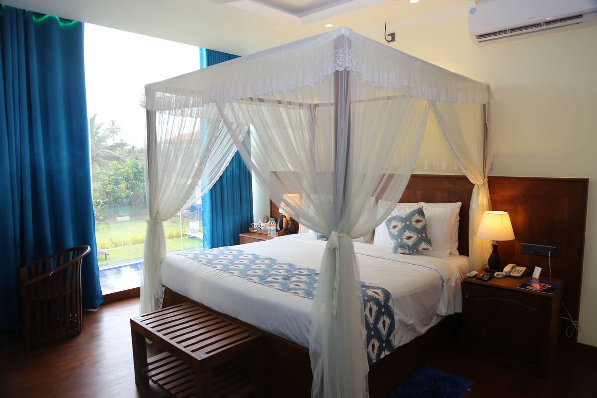 A four-poster canopy bed with a view of a garden, a wooden bench, a nightstand with a lamp, and a telephone.
