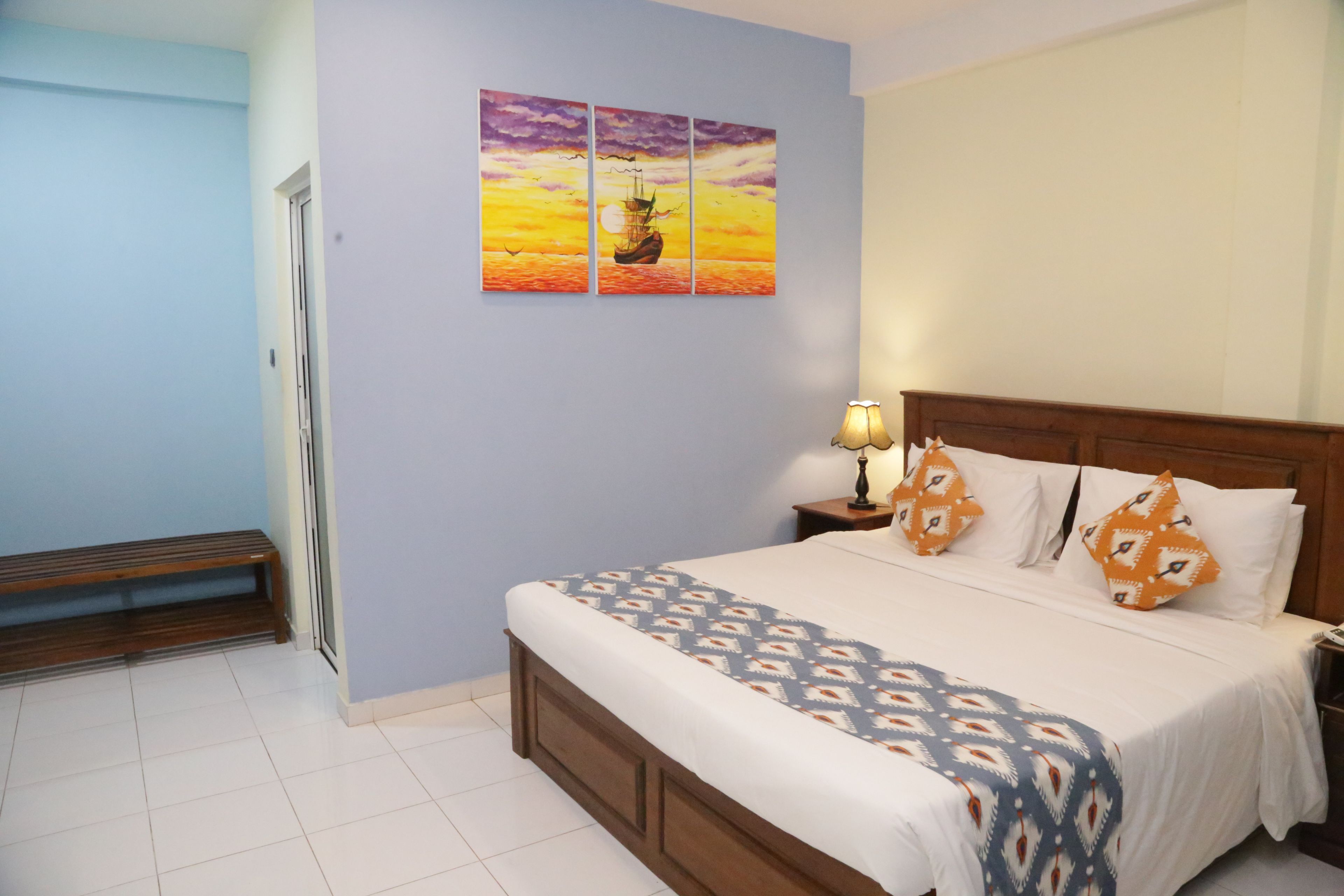 A hotel room with a bed, a nightstand, a lamp, and a wall with a sunset painting.