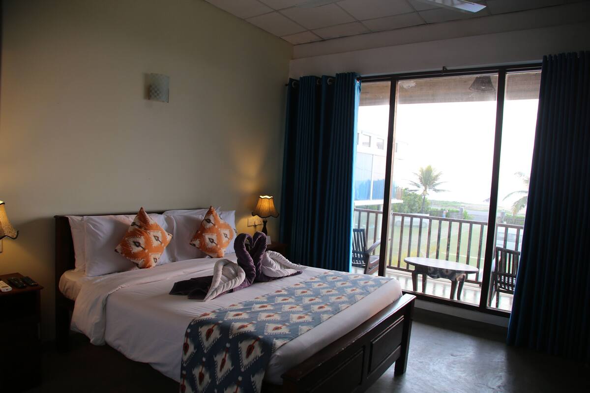 A hotel room with a bed, pillows, a bedside table with a lamp, a window with curtains, and a balcony with a table and chairs.