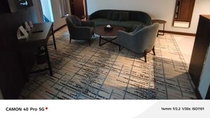 Ein modernes Wohnzimmer mit einer Couch, zwei Sesseln, einem Couchtisch und einem Flachbildfernseher. Der Raum verfügt über einen gemusterten Teppich und ein großes Fenster mit Vorhängen.