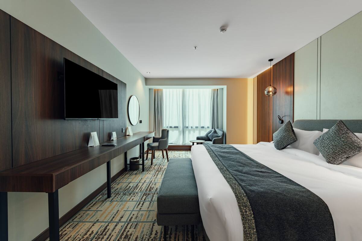A modern hotel room with a large bed, a desk, a flat-screen TV mounted on the wall, and a large window with curtains.