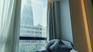 Ein Zimmer mit Blick auf eine Stadtlandschaft durch ein Fenster, ein grauer Sessel mit einem strukturierten Kissen und ein zur Seite gezogen Vorhang.