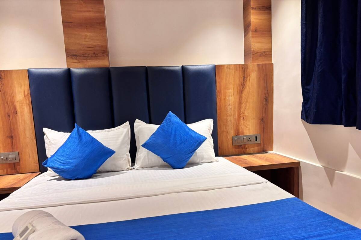 Una camera d'albergo con un letto, cuscini bianchi e azzurri, copripiumino blu, una testiera in legno e un comodino anch'esso in legno con una presa di corrente.