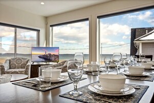 Un comedor con una mesa lista para una comida, ventanas amplias con vista al mar y un televisor mostrando una escena de atardecer.