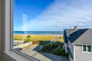 Vista costera desde una ventana que da a una playa, una zona con césped y un edificio con techo inclinado.
