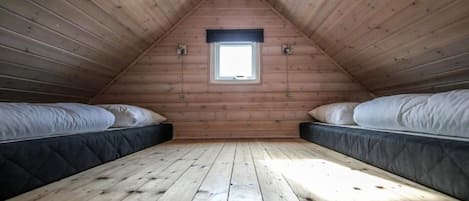 Um quarto de sótão pequeno com paredes de madeira, teto inclinado e uma única janela. Há duas camas com colchões e travesseiros.