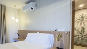 A hotel room with a bed, bedside tables, a wall-mounted air conditioner, a nightstand with a lamp, and a decorative wall piece.