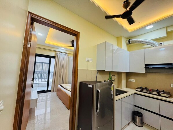 Une cuisine compacte équipée d’un réfrigérateur, d’une cuisinière et de placards. On y trouve une petite poubelle et un ventilateur de plafond. Une porte donne accès à un balcon où se trouve un lit.