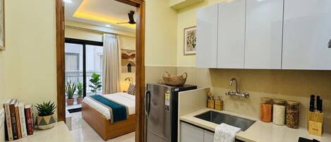 Habitación de hotel compacta con kitchenette, una cama con manta azul, una pequeña mesa de comedor y una ventana con cortinas.