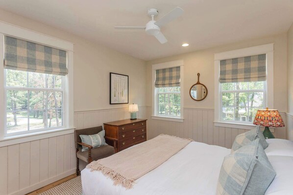 A bedroom with a bed, a dresser, a chair, a ceiling fan, and two windows with checkered blinds.