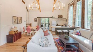 A spacious living room with a large white sofa, a wooden coffee table, and a colorful rug. There is a kitchen area with a window and a dining table with chairs.