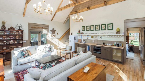 Un salón amplio con un sofá grande, una mesa de centro y una mesa de comedor de madera. Cuenta con una zona de cocina con encimera y nevera. La habitación tiene vigas de madera en el techo y un candelabro.