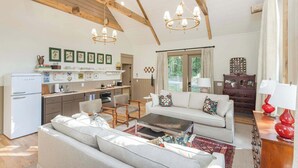 Amplia sala de estar con sofá, mesa de centro y mesa de comedor. Cuenta con una cocina equipada con refrigerador y fregadero. La habitación tiene vigas de madera en el techo y un ventanal grande.