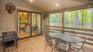 Un espacio al aire libre techado con una terraza de madera, una mesa redonda con sillas, una parrilla y vista a un bosque.