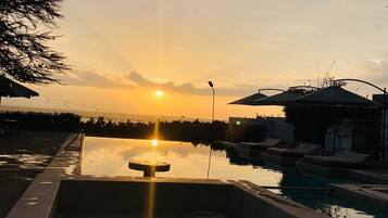 A swimming pool with a sunken seating area, surrounded by lounge chairs and umbrellas, with a sunset in the background.