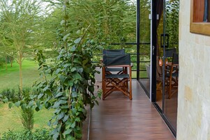 A balcony with a wooden floor, a table with chairs, and a view of a garden with trees and shrubs.