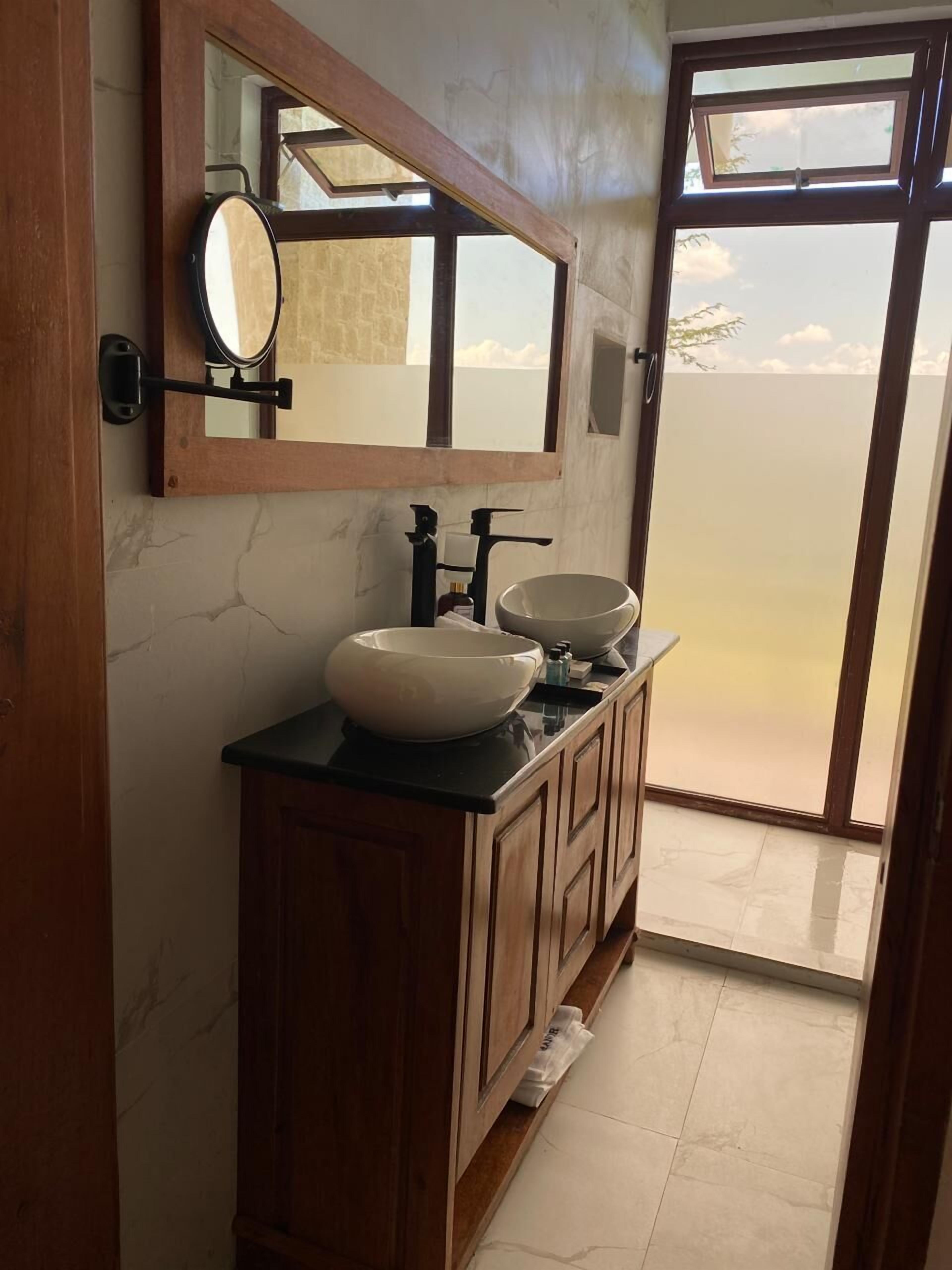 Ein Badezimmer mit einem Holzbadezimmermöbel, zwei Waschbecken, einem großen Spiegel und Blick durch eine Glastür.