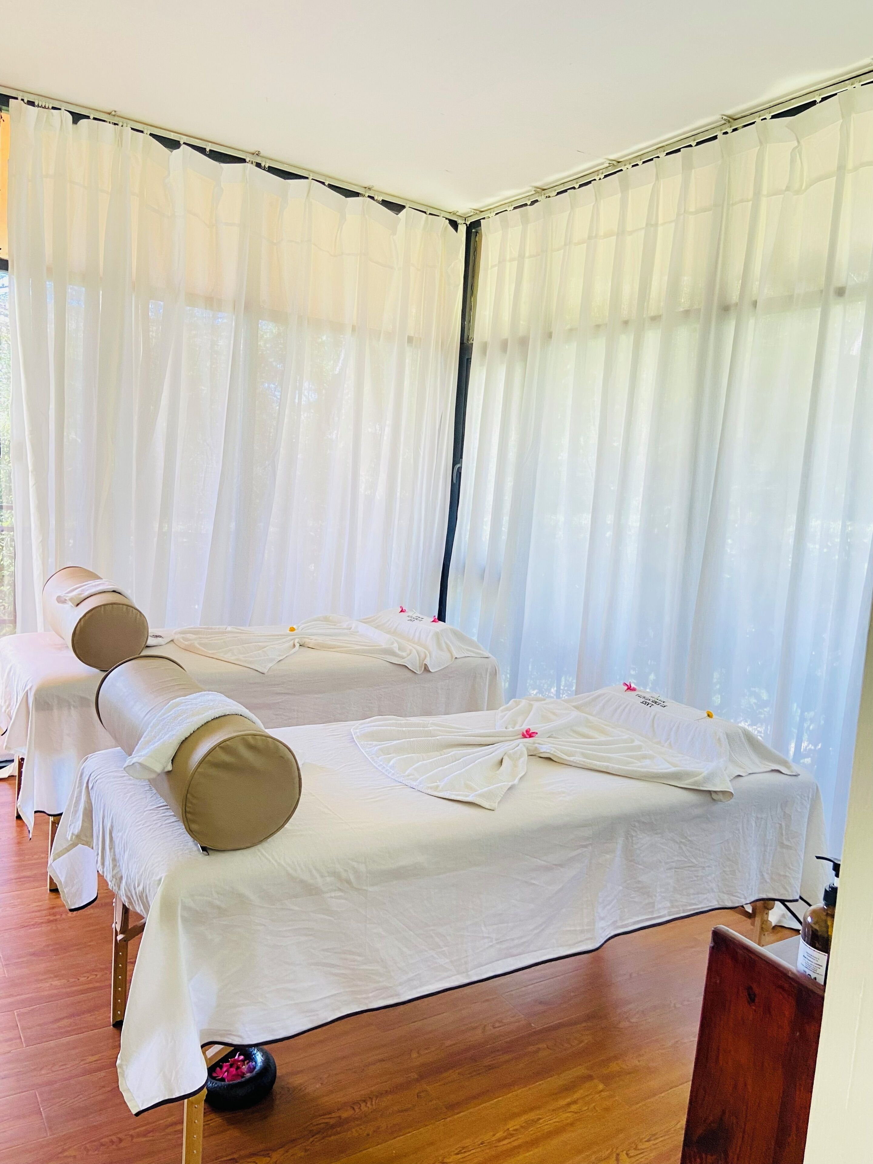 A spa room with two massage beds, a wooden floor, and sheer curtains.