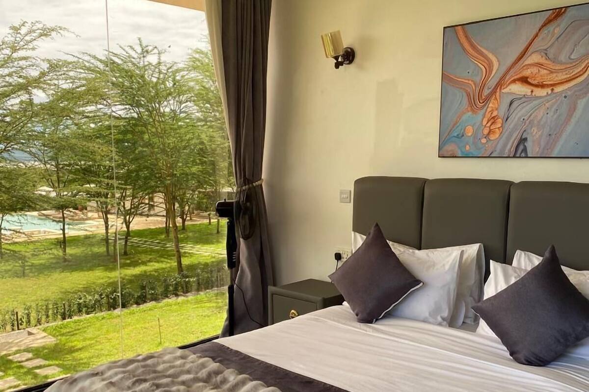 A hotel room with a large bed, a view of a grassy area and trees, and a painting on the wall.