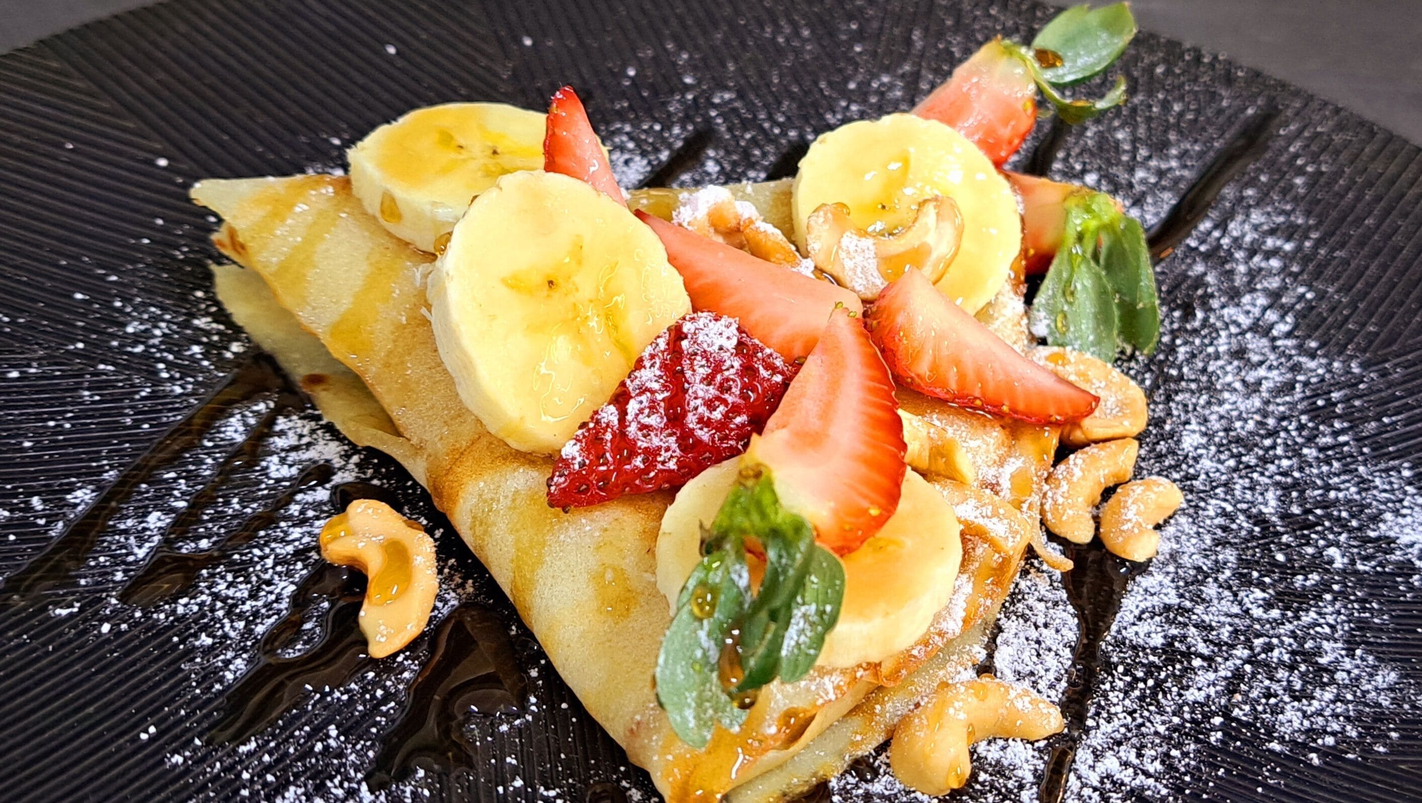 Un plato de postre con un crepe cubierto de rodajas de banana, fresas y un poco de azúcar glas.