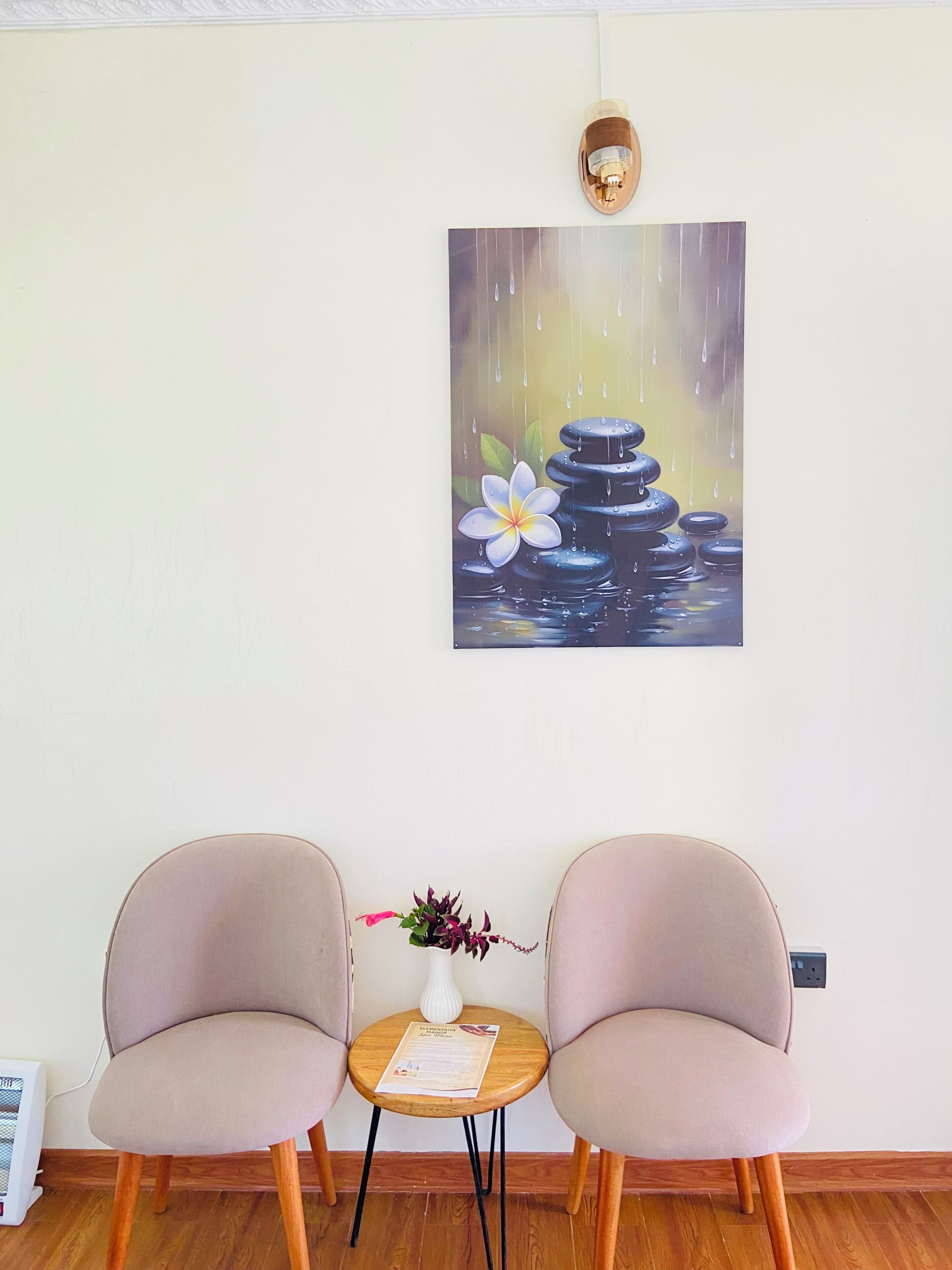 A room with two chairs, a small table with a vase, and a painting of a water feature.