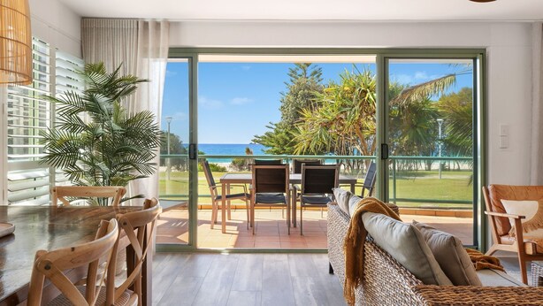 Una habitación moderna con un amplio ventanal corredero que da a un balcón con vista al océano. Hay una mesa y sillas de madera, un sofá de fibras naturales y una planta en maceta.