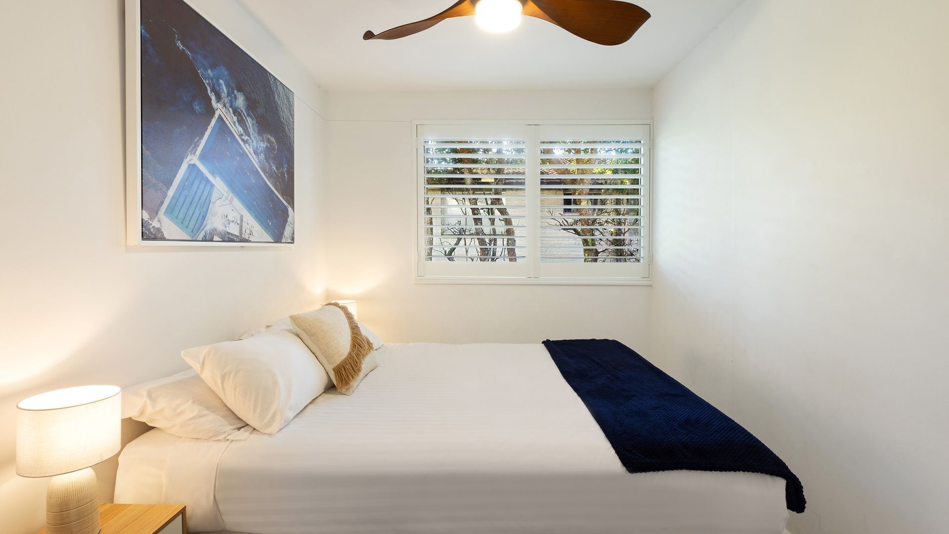 A bedroom with a bed, a nightstand, a lamp, a ceiling fan, a window with blinds, and a wall-mounted picture.