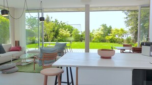 Un salon moderne doté d’une grande porte vitrée donnant sur un jardin verdoyant. On y trouve un canapé blanc, une table basse en verre et une table à manger en bois avec des chaises.