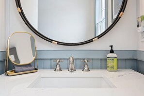 A bathroom with a white sink, three faucets, a gold-framed mirror, a black circular object above the mirror, and a bottle of hand soap on the counter. - Steps to Beach & Downtown OGT - Screened in Porch (Ogunquit)