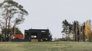 A modern, black building with a flat roof and large windows, surrounded by trees and a grassy field.