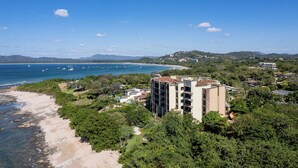 Un hotel frente al mar con varios pisos, rodeado de exuberante vegetación y una playa.