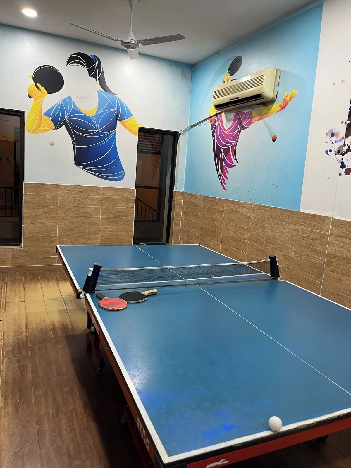 Una mesa de ping pong con dos paletas y una pelota, un ventilador de techo y una pared con un mural de una persona jugando ping pong.