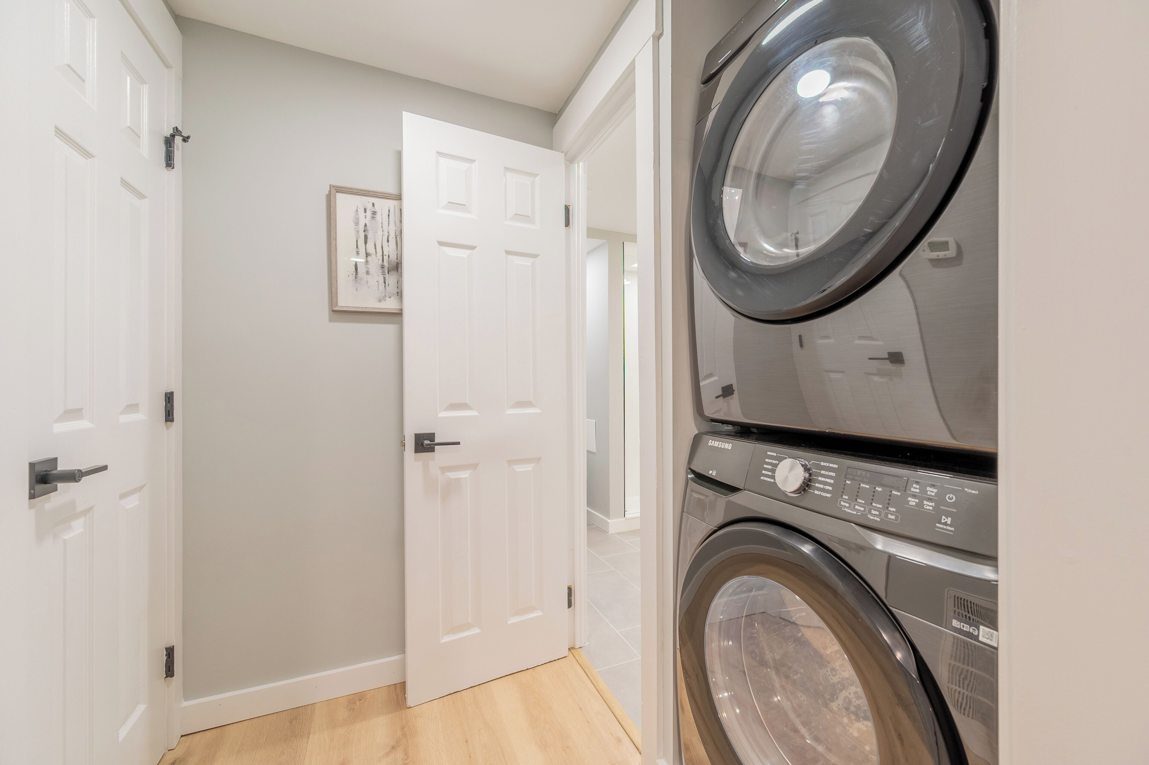 Une buanderie équipée d’un lave-linge et d’un sèche-linge superposés, d’une porte blanche et d’un tableau encadré accroché au mur.