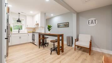 A modern kitchen with white cabinets, a wooden dining table with stools, a trash can, and a wall-mounted sign that says "EAT".