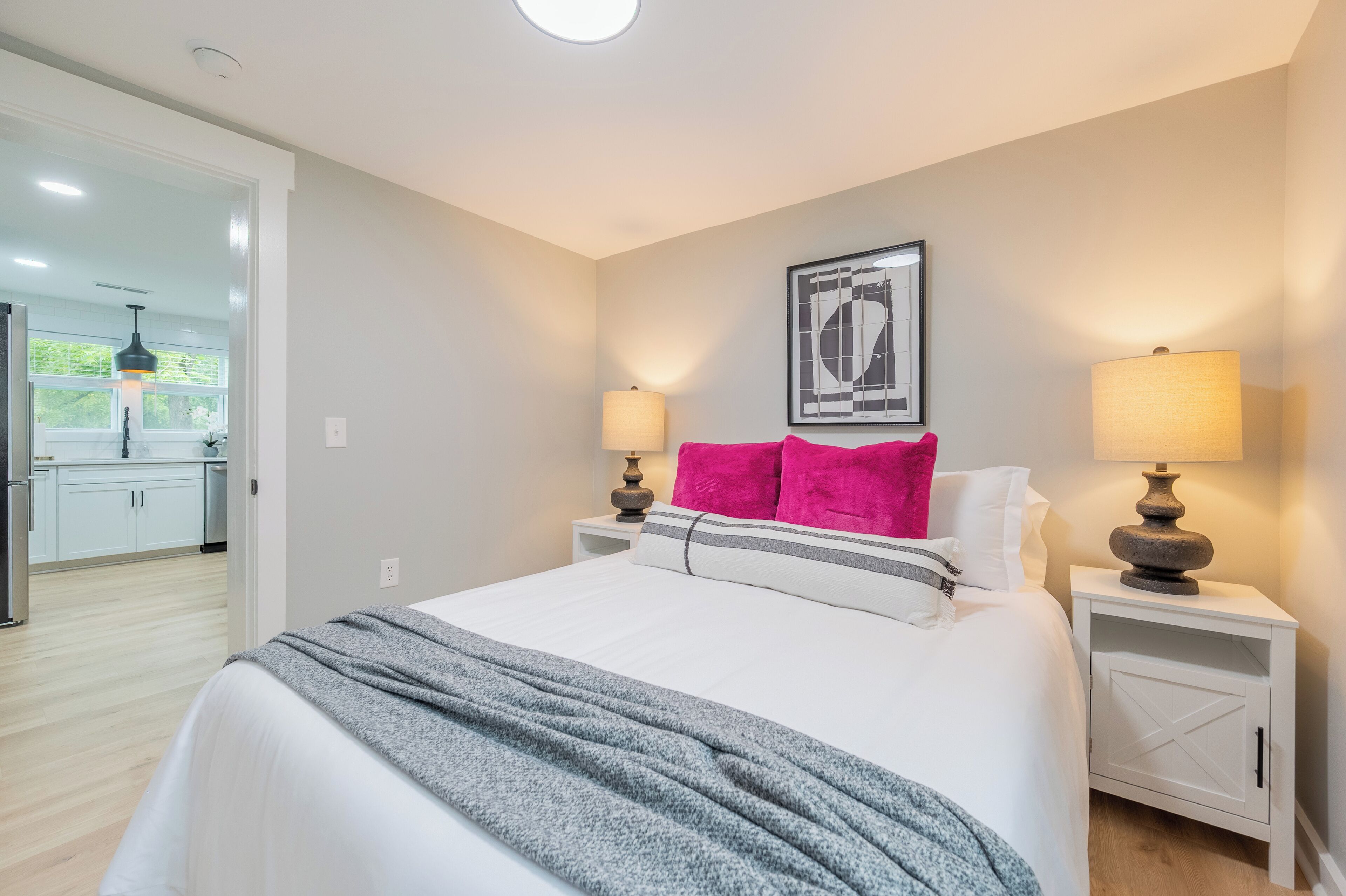 A bedroom with a bed, two bedside tables with lamps, a framed artwork above the bed, and a view into the kitchen.
