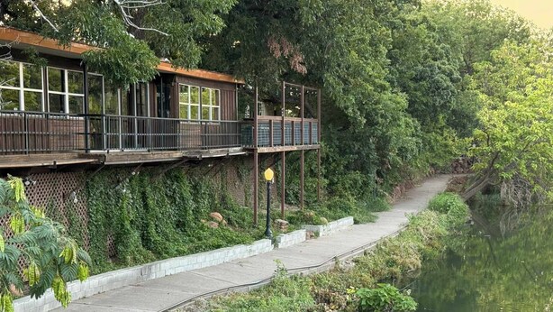 A riverside walkway with a wooden deck and railing, surrounded by lush greenery and a calm river.