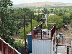 Une vue depuis un balcon doté d’une balustrade en métal, donnant sur un paysage verdoyant avec des arbres et des collines.