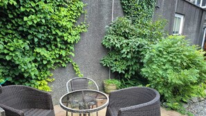 Un espace patio aménagé avec des meubles en osier et une table en verre, entouré d’une végétation luxuriante et d’un mur en pierre.
